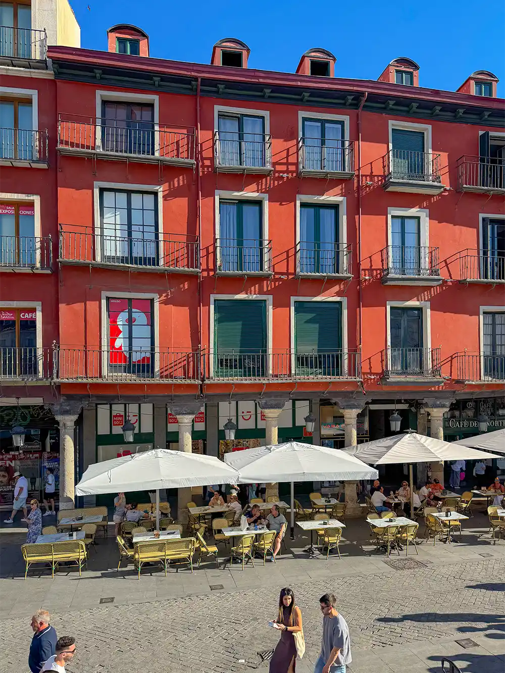 Valladolid-Plaza-Mayor An einem sonnigen Tag sitzen Menschen an den Tischen eines Straßencafés unter einem roten Gebäude mit Balkonen.