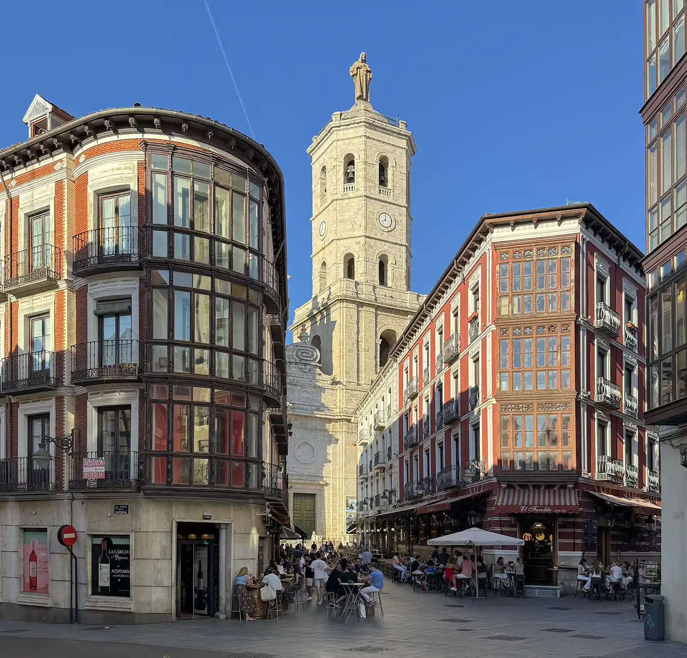 Valladolid Menschen sitzen in Straßencafés zwischen bunten Gebäuden, im Hintergrund ragt ein hoher Kirchturm vor blauem Himmel empor.