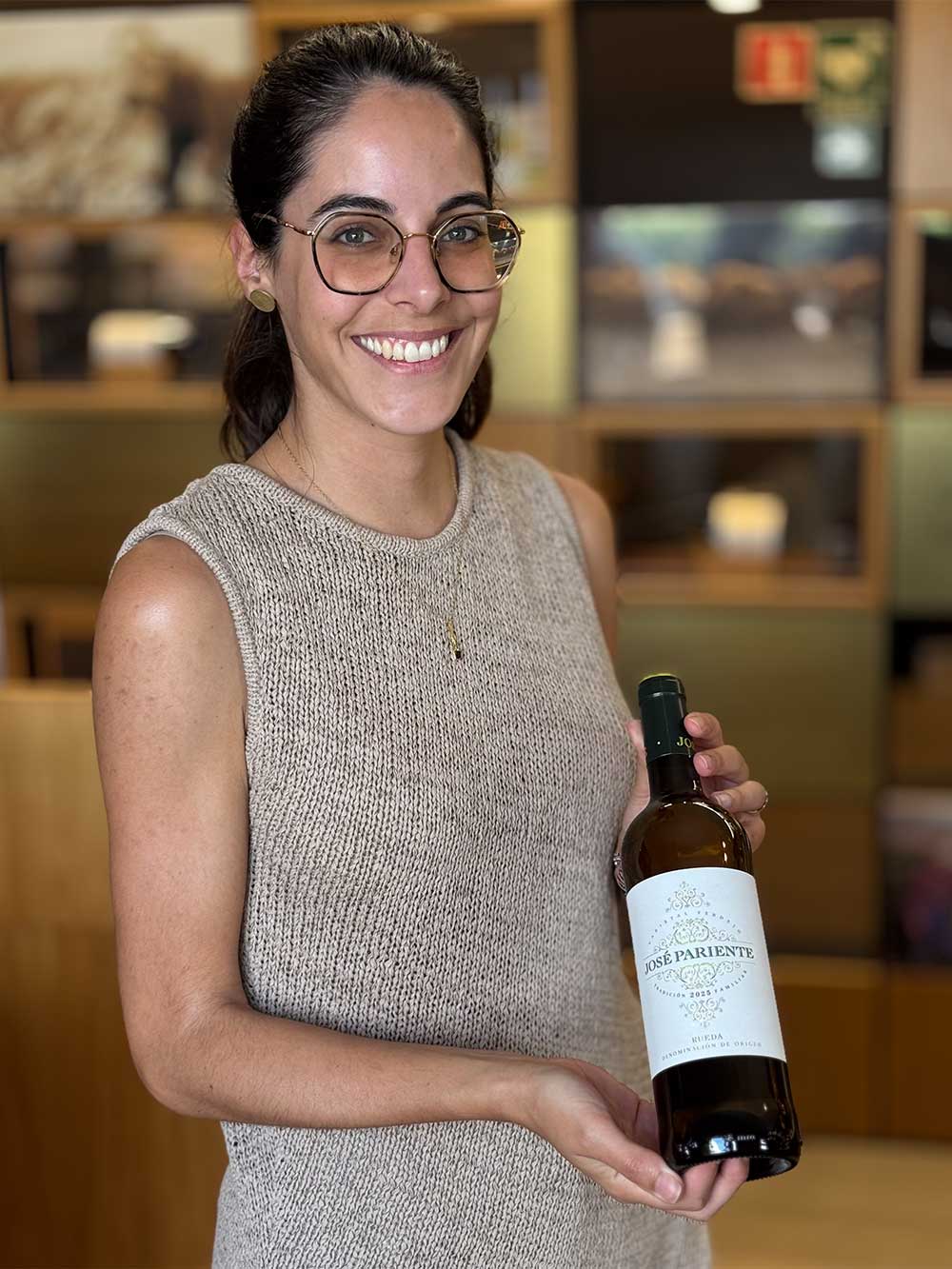 Verdejo-Rueda-Jose-Pariente Eine Frau mit Brille, die lächelt und eine Flasche José Pariente-Weißwein in einem Innenraum in der Hand hält.