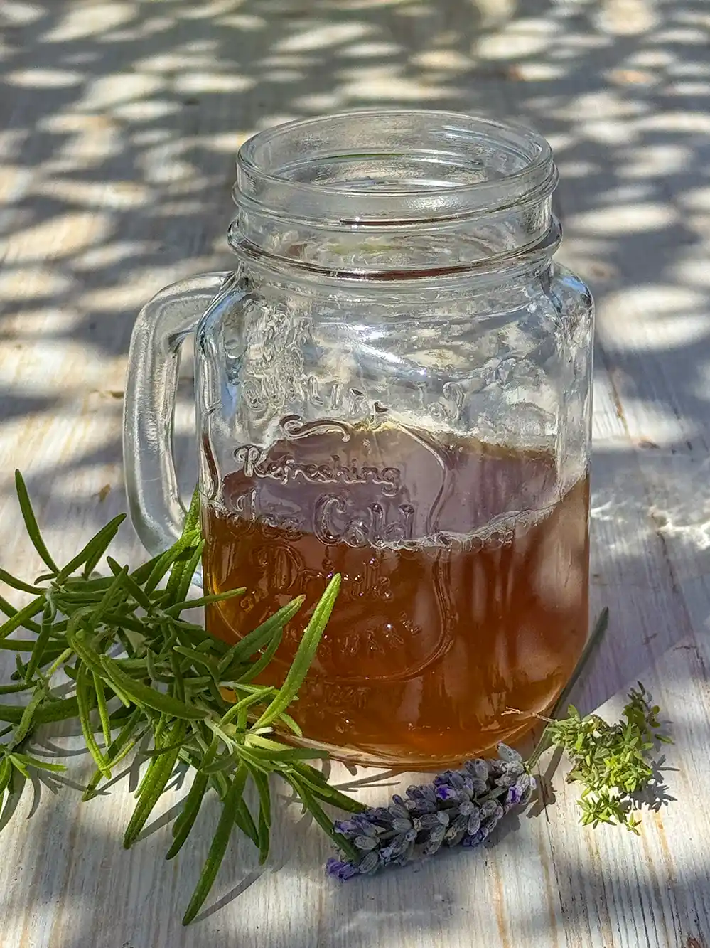 Ein Einmachglas mit Eistee auf einem Holztisch, daneben Rosmarin- und Lavendelzweige im Sonnenlicht.