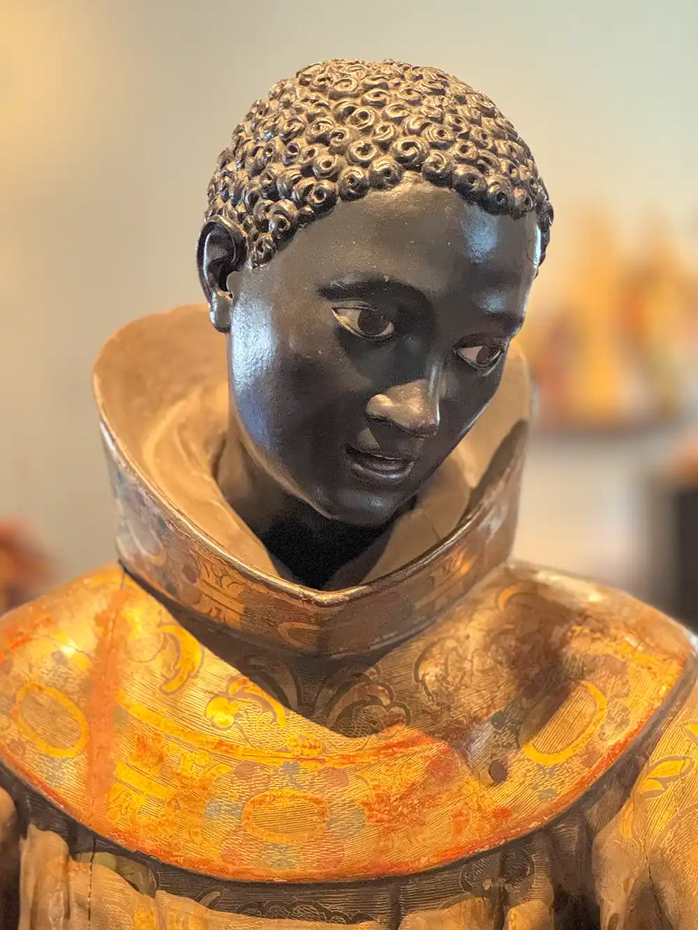 Benedikt-der-Mohr-Palermo Holzstatue einer Person mit lockigem Haar, dunklem Gesicht, verziertem Kragen und einem Gewand mit goldenen Mustern.