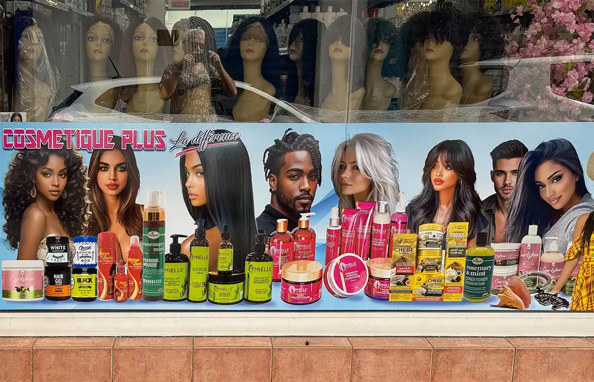 Hautfarben-Martinique-Schoenheitssalon Schaufenster, in dem Haarpflegeprodukte ausgestellt sind, hinter denen Plakate mit verschiedenen Models zu sehen sind, die unterschiedliche Frisuren präsentieren.