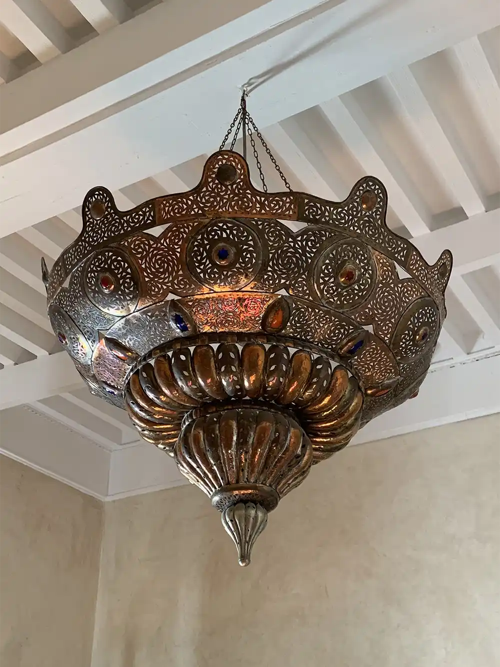 Kolonialstil-marokkanische-Lampe Lampe aus Bronze mit farbigen Glassteinen, der von einer weißen Balkendecke herabhängt.