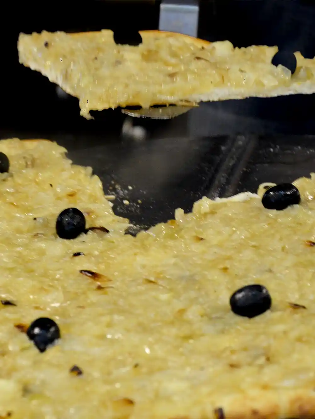 Pissaladiere-Nizza-Zwiebel Eine Nahaufnahme eines Zwiebelnkuchens mit schwarzen Oliven, der gerade in Stücke geschnitten wird, wobei ein Stück angehoben wird.