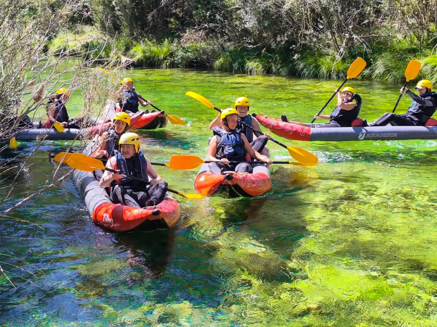 Alto-Tajo-Kajak-wandern Eine Gruppe von Menschen mit Helmen paddelt in aufblasbaren Kajaks auf einem klaren, grünen Fluss, der von üppigen Bäumen umgeben ist.