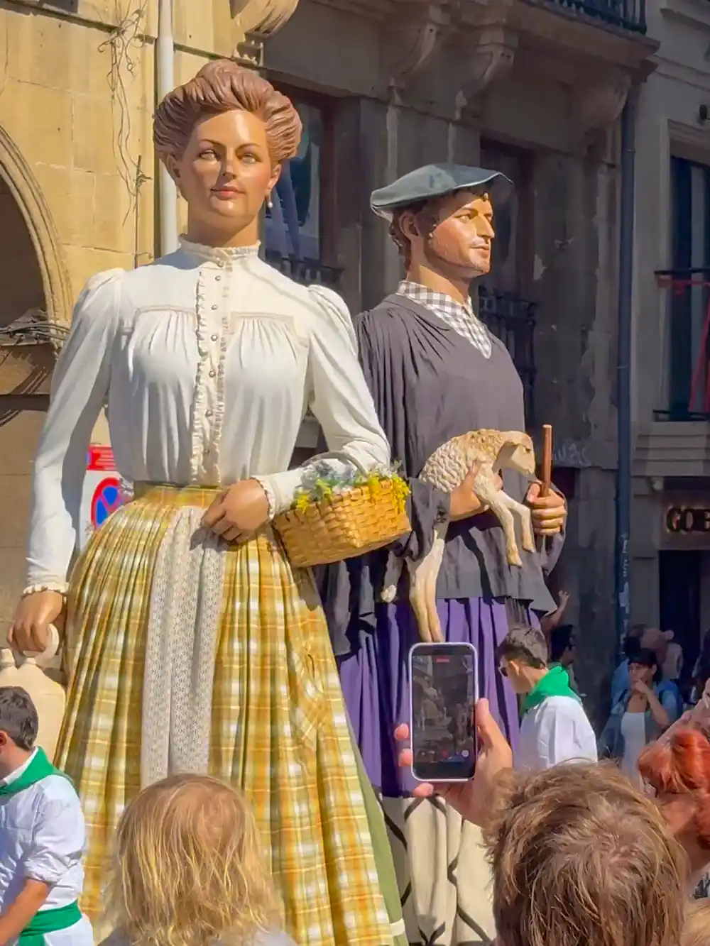 Aste-Nagusia-Bilbao-Giganten-Ehepaar Zwei große, kostümierte Figuren bei einem Umzug: Die eine hält einen Korb in der Hand, die andere ein Lamm, während die Menschen zuschauen.