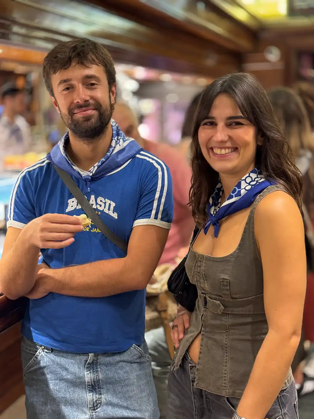 Aste-Nagusia-Bilbao-Paar Zwei Menschen, die auf einem Markthallenmarkt lächeln; beide tragen blaue Halstücher; einer hält etwas zu essen in der Hand.