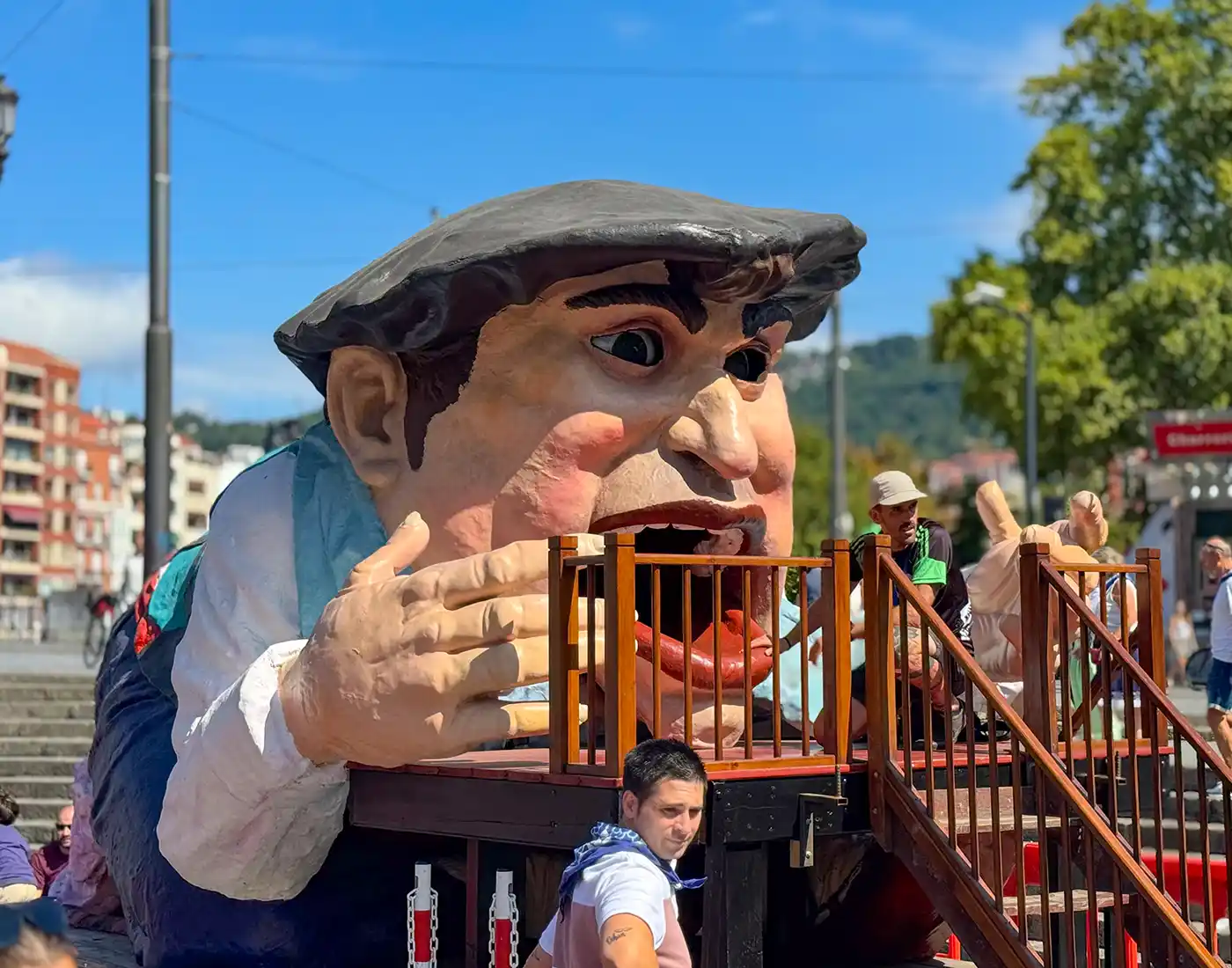 Aste-Nagusia-Gargantua Ein Festwagen mit einem riesigen Figurenkopf, umgeben von Menschen im Freien an einem sonnigen Tag.