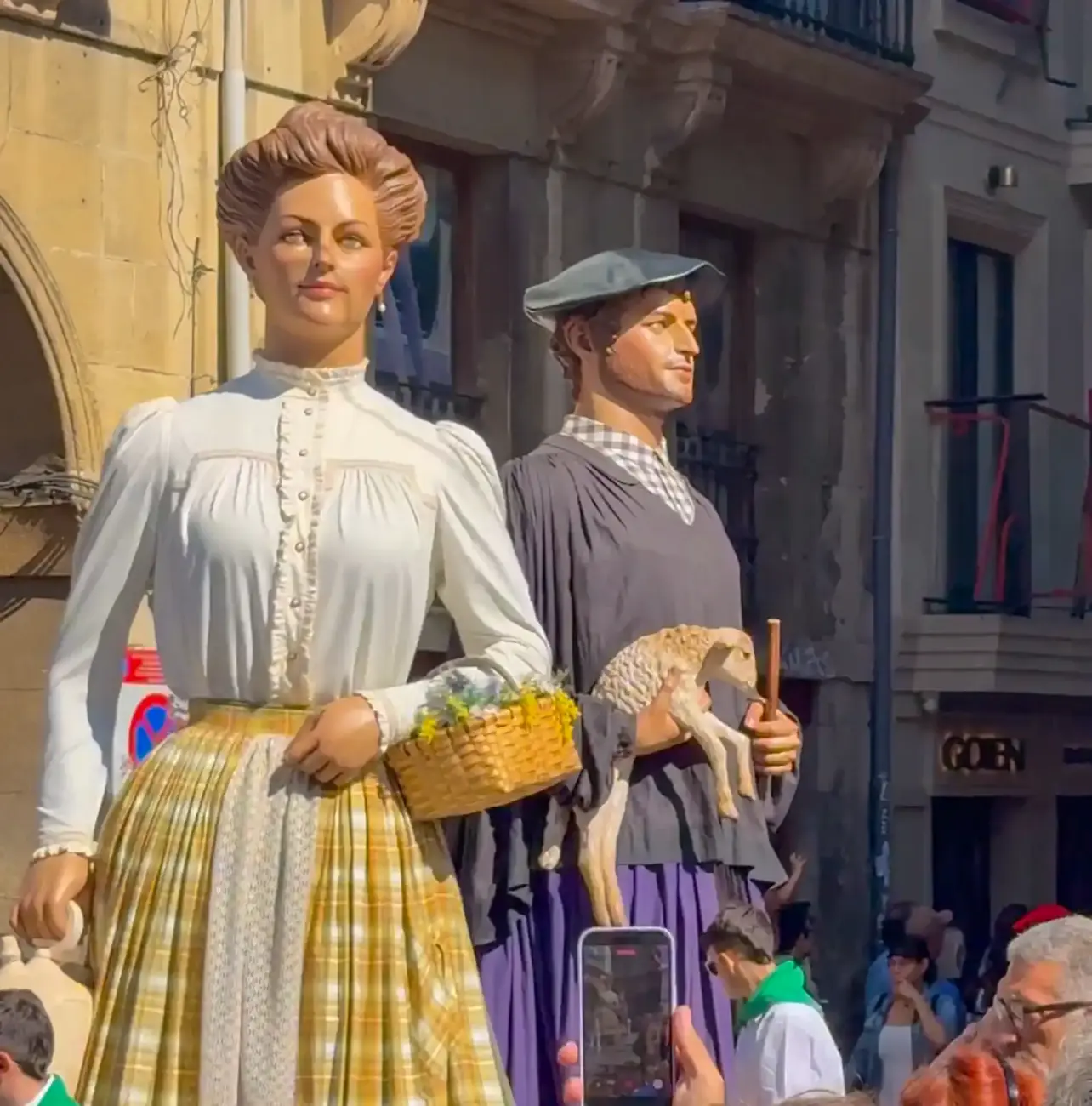Zwei große, traditionell gekleidete Festfiguren, von denen eine einen Korb hält und die andere ein Lamm trägt, bei einem Straßenumzug.