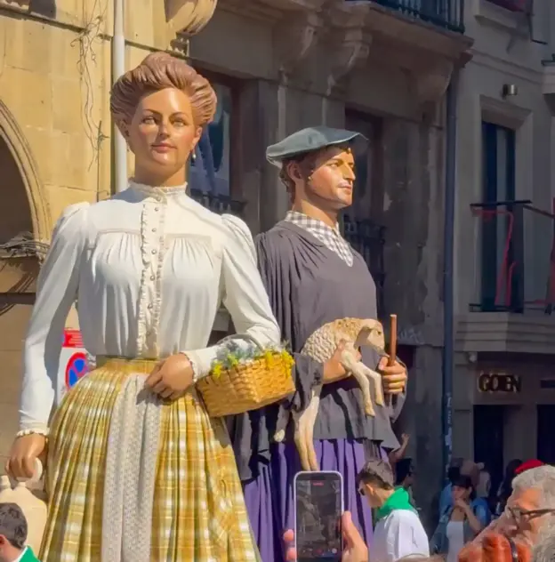 Zwei große, traditionell gekleidete Festfiguren, von denen eine einen Korb hält und die andere ein Lamm trägt, bei einem Straßenumzug.