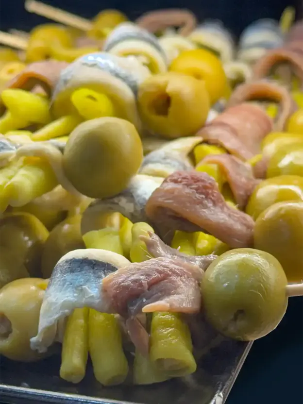 Nahaufnahme von Spießen mit grünen Oliven, Sardellen und eingelegten Paprikaschoten, die auf einem Tablett angerichtet sind.