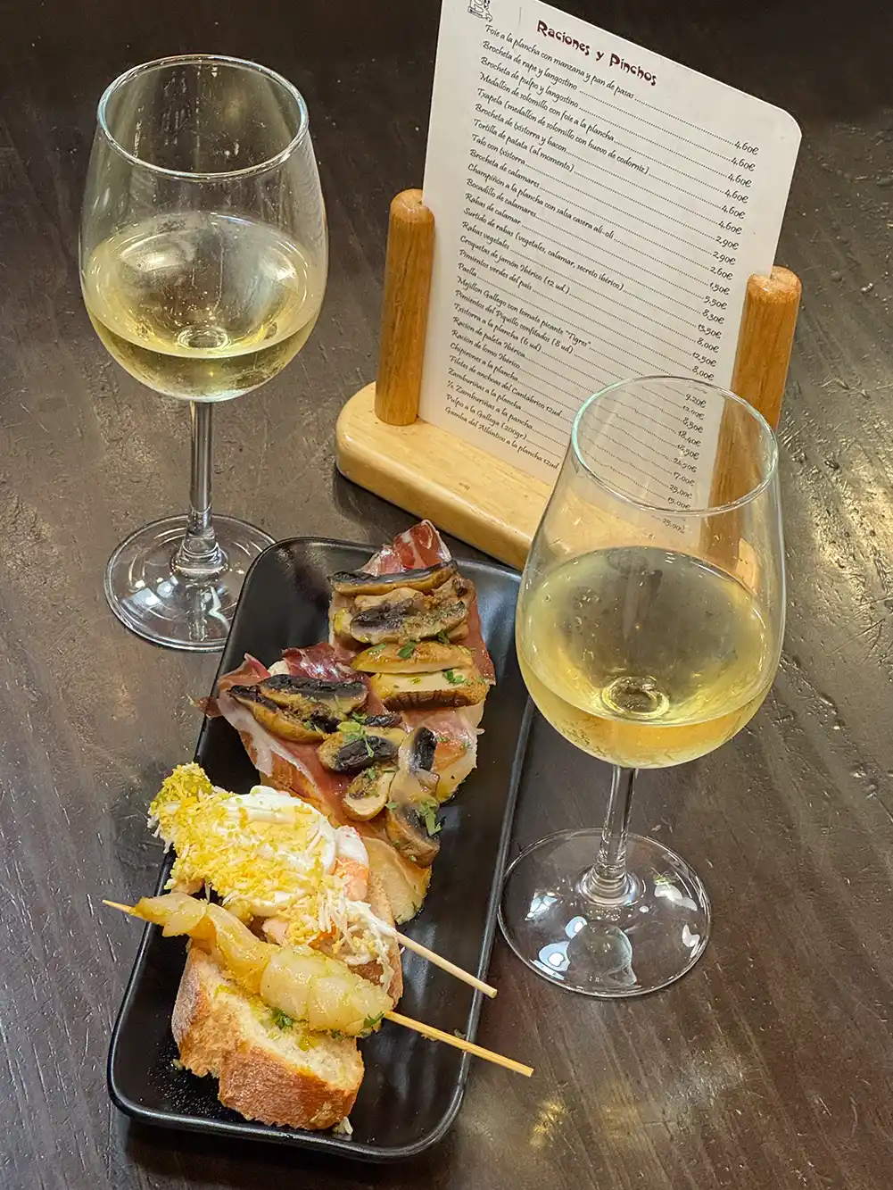 Zwei Gläser Weißwein mit drei verschiedenen Tapas auf einem schwarzen Teller, im Hintergrund die Speisekarte auf einem dunklen Tisch.