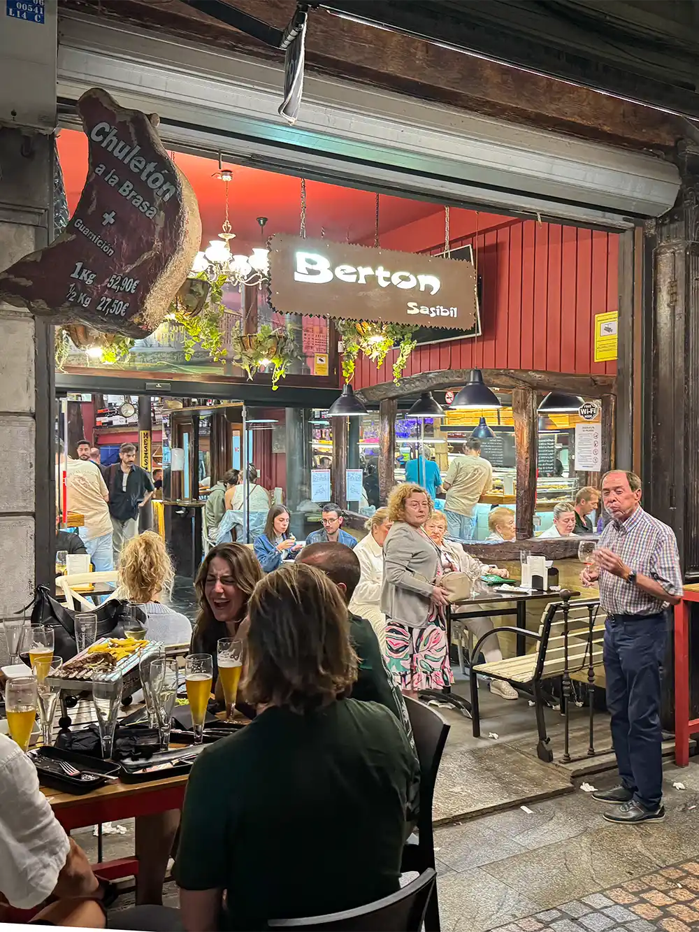 Menschen, die abends in einem Restaurant im Freien essen und sich unterhalten, im Hintergrund sind Berton-Schilder zu sehen.