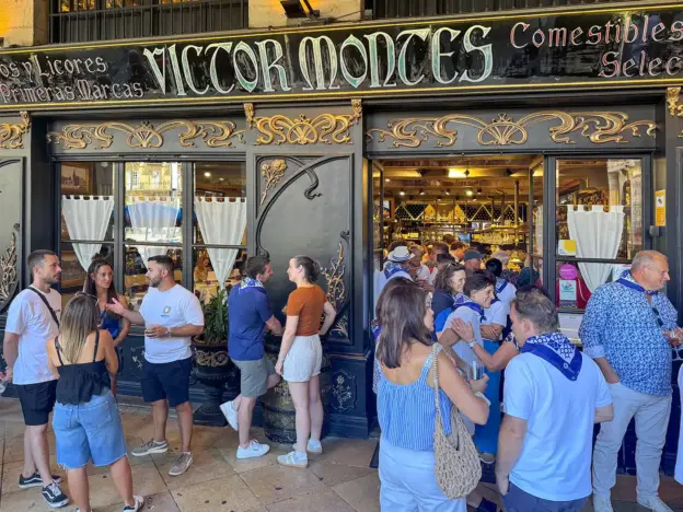 An einem sonnigen Tag versammeln sich vor dem Eingang des Restaurants „Victor Montes“ Menschen und unterhalten sich.