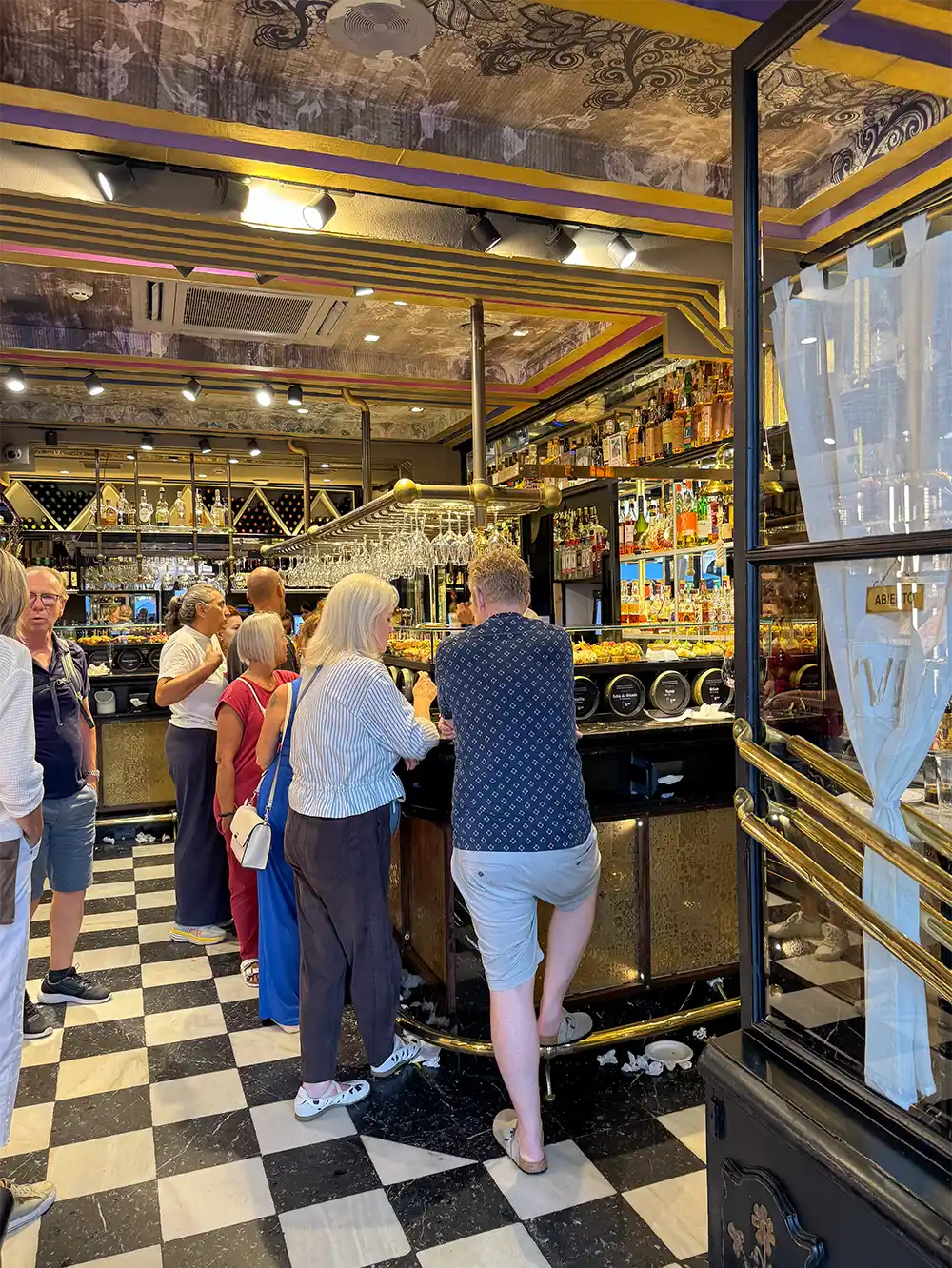 Die Gäste stehen an einer stilvollen Bar mit kariertem Boden, Regalen voller Flaschen und dekorativen Details an der Decke.