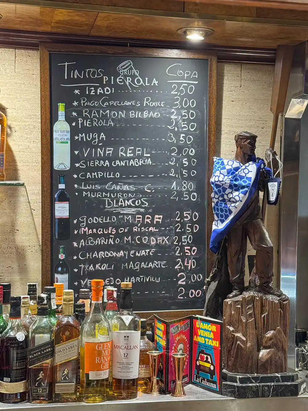 Bilbao-Pintxo-Bars-Amaya Eine Weinkarte auf einer Kreidetafel über der Bar, daneben Spirituosenflaschen und eine Holzstatue auf der Theke.