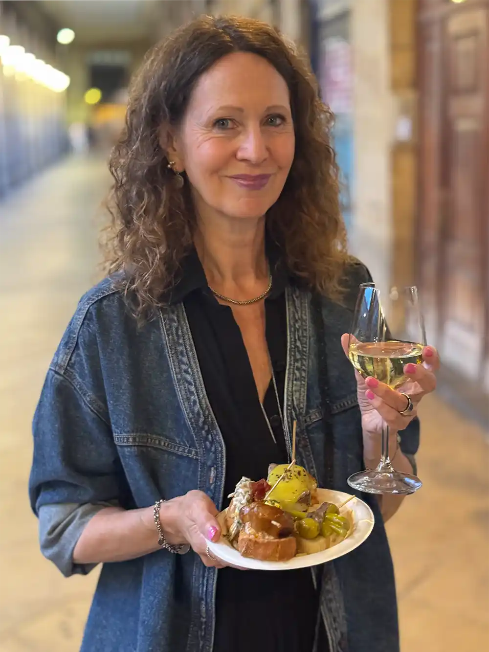 Bilbao-Pintxo-Bars-Tour Eine Frau mit lockigem Haar hält einen Teller mit Essen und ein Glas Weißwein in der Hand und lächelt in einem Flur.