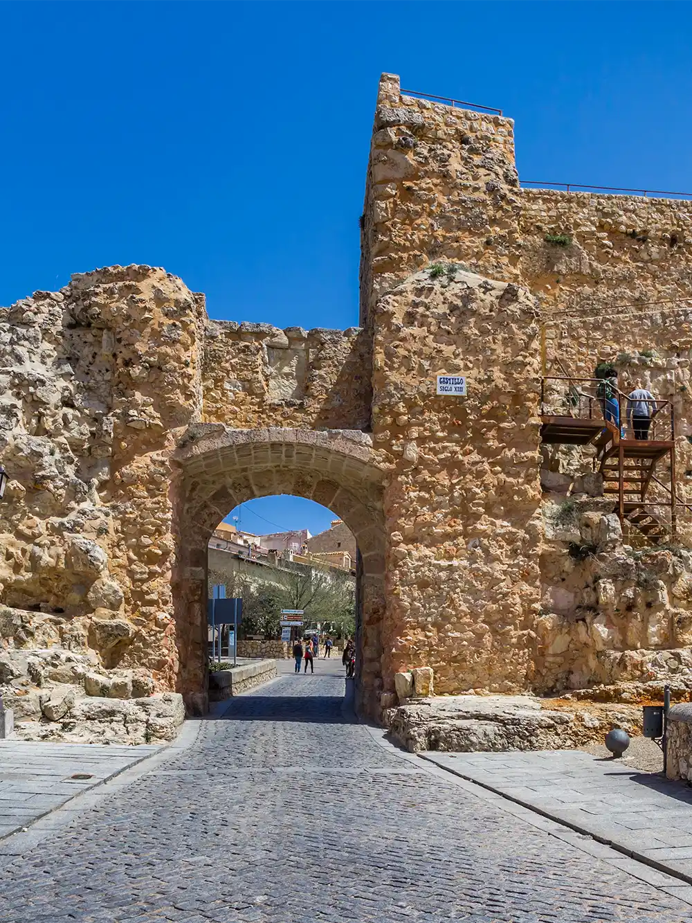 Cuenca-Alcazar-Ruinen Ein steinernes Burgtor mit gewölbtem Eingang, alten Mauern und Stufen unter strahlend blauem Himmel.