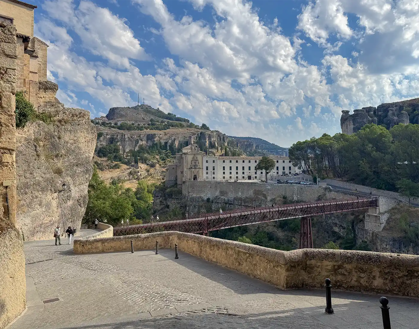 Cuenca-Blick-Parador-Bruecke Eine Steinbrücke überspannt eine tiefe Schlucht, hinter der sich historische Gebäude und grüne Hügel vor einem teilweise bewölkten Himmel erstrecken.