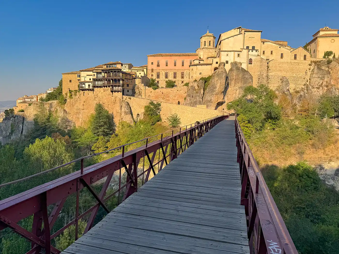 Cuenca-Bruecke Eine Fußgängerbrücke aus Holz, die zu alten Steinhäusern auf einem felsigen Hügel unter strahlend blauem Himmel führt.