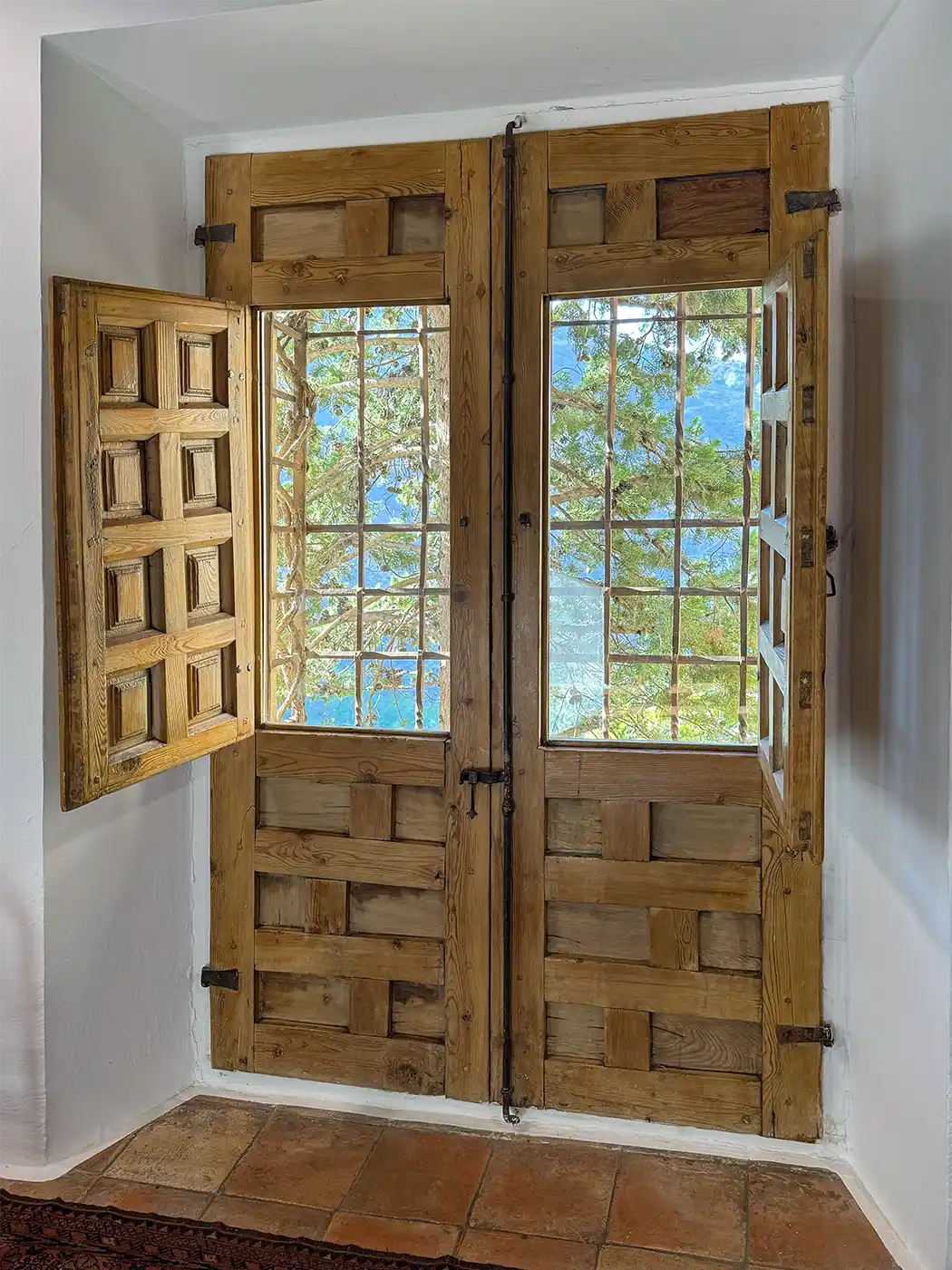 Cuenca-Casas-Colgadas-innen Ein Holzfenster mit Eisenstangen, auf einer Seite offen, durch das man Bäume und blaues Wasser draußen sehen kann.