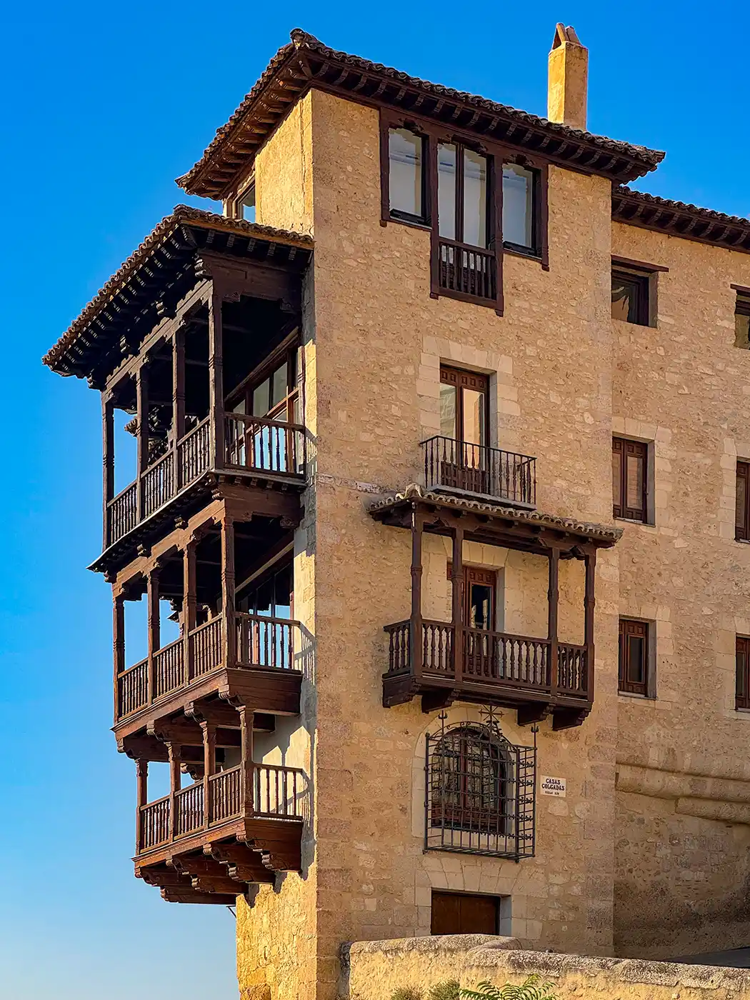 Cuenca-Casas-Colgadas Ein Steingebäude mit nach außen ragenden Balkonen aus dunklem Holz, vor dem Hintergrund eines strahlend blauen Himmels.