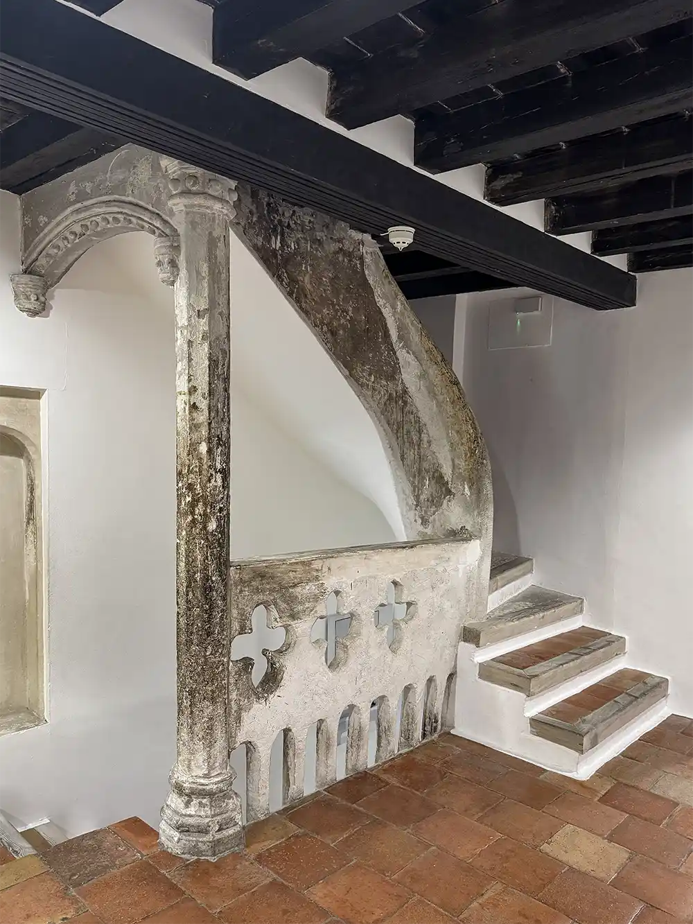 Cuenca-Casas-colgadas-innen Eine kleine mittelalterliche Steintreppe mit kunstvollem Geländer neben einer modernen Treppe und einer dunklen Holzdecke.