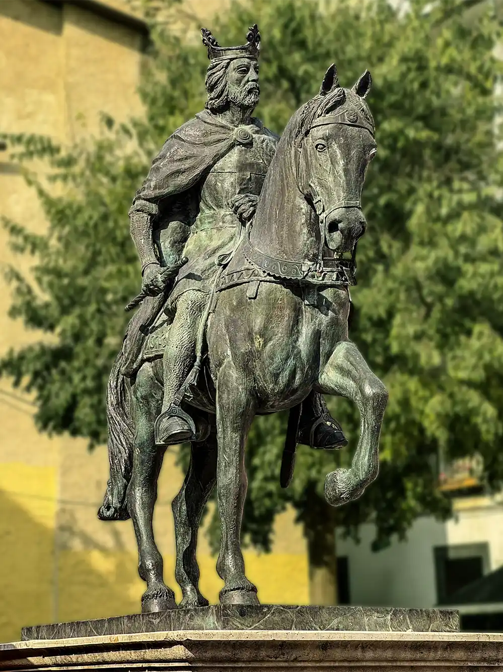 Cuenca-Denkmal-Alfons-VIII Bronzestatue eines gekrönten Mannes in Rüstung auf einem Pferd, im Hintergrund sind Bäume und Gebäude zu sehen.