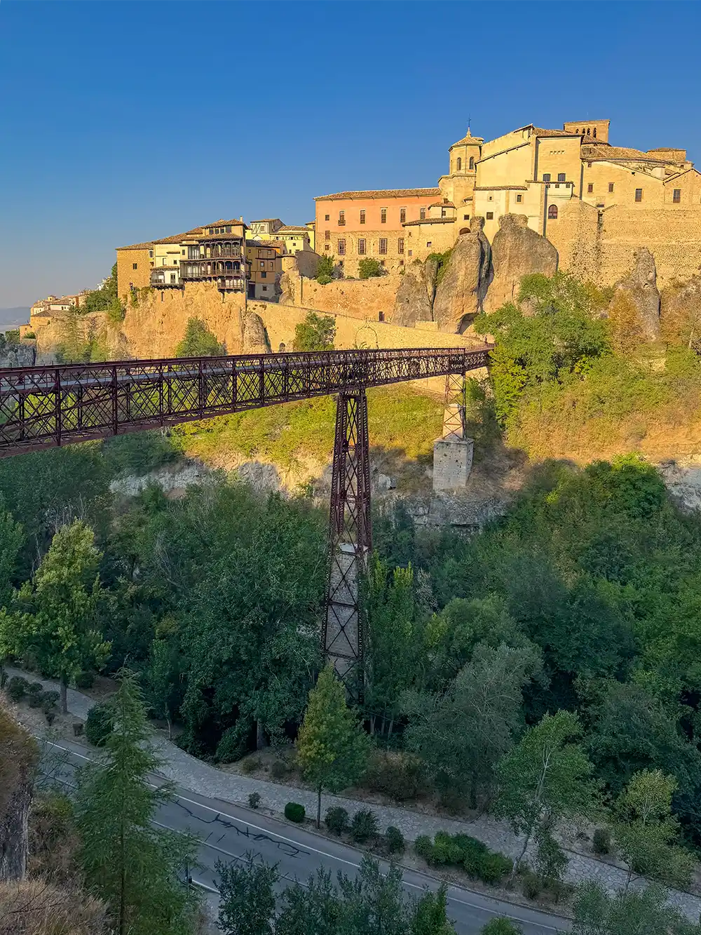 Cuenca-Eisen-Bruecke Eine Eisenbrücke überspannt eine grüne Schlucht, über der sich unter einem strahlend blauen Himmel historische Gebäude auf einem felsigen Klippenvorsprung erheben.