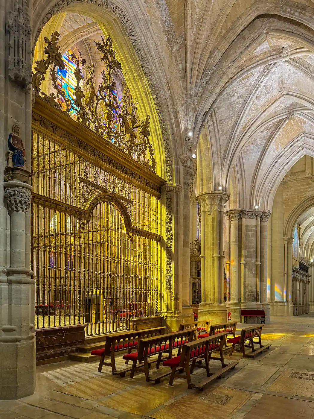 Cuenca-Kathedrale-Lichtstimmung In einer prächtigen Kathedrale mit Gewölbedecke stehen Reihen von Holzbänken vor einem reich verzierten, goldenen Altar.