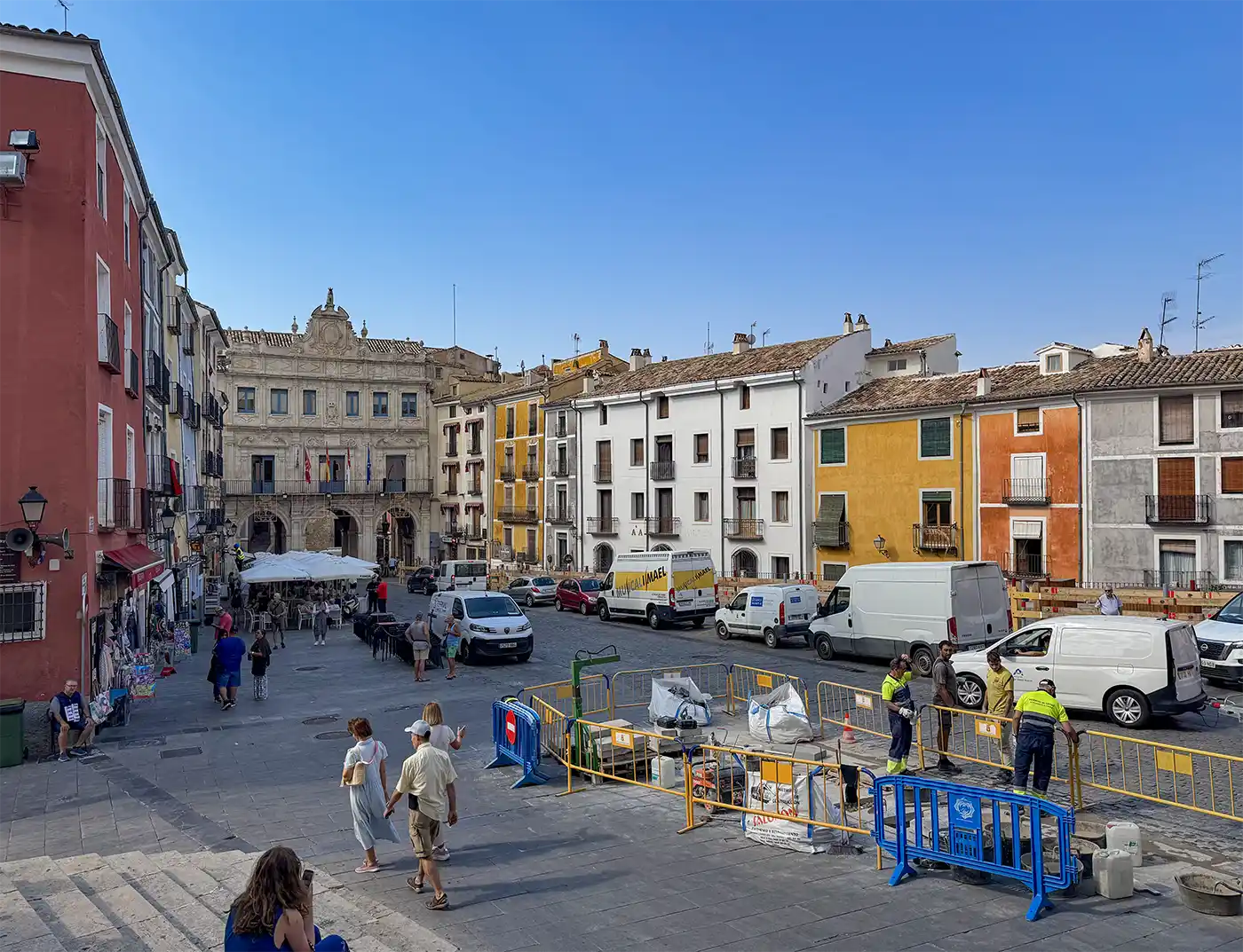 Cuenca-Plaza-Mayor-San-Mateo Menschen spazieren über einen farbenfrohen europäischen Platz; Arbeiter und Transporter stehen in der Nähe von Absperrungen, im Hintergrund sind Gebäude zu sehen.