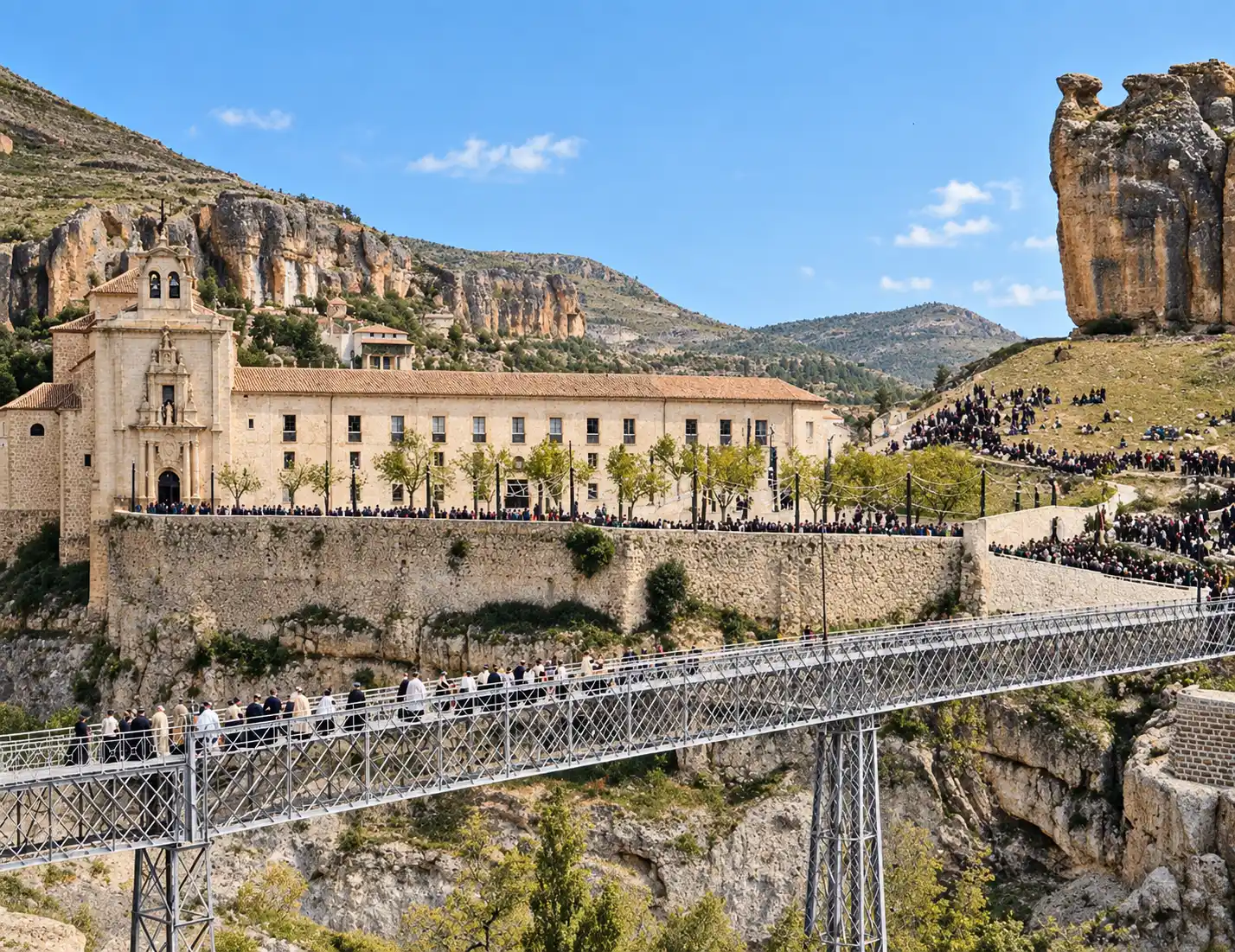 Cuenca-Puente-de-San-Pablo-1903 Ein großes Steingebäude, Menschenmengen auf einer Brücke und an einem Hang, im Hintergrund felsige Klippen und blauer Himmel.
