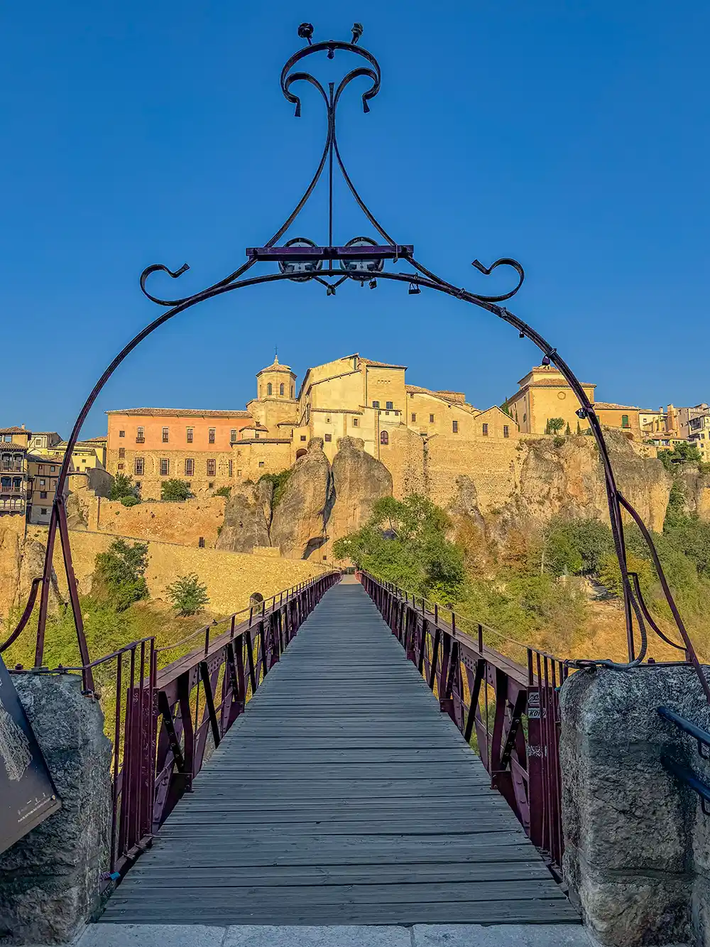 Cuenca-Puente-de-San-Pablo Eine hölzerne Fußgängerbrücke führt zu historischen Gebäuden auf einer felsigen Klippe unter strahlend blauem Himmel.