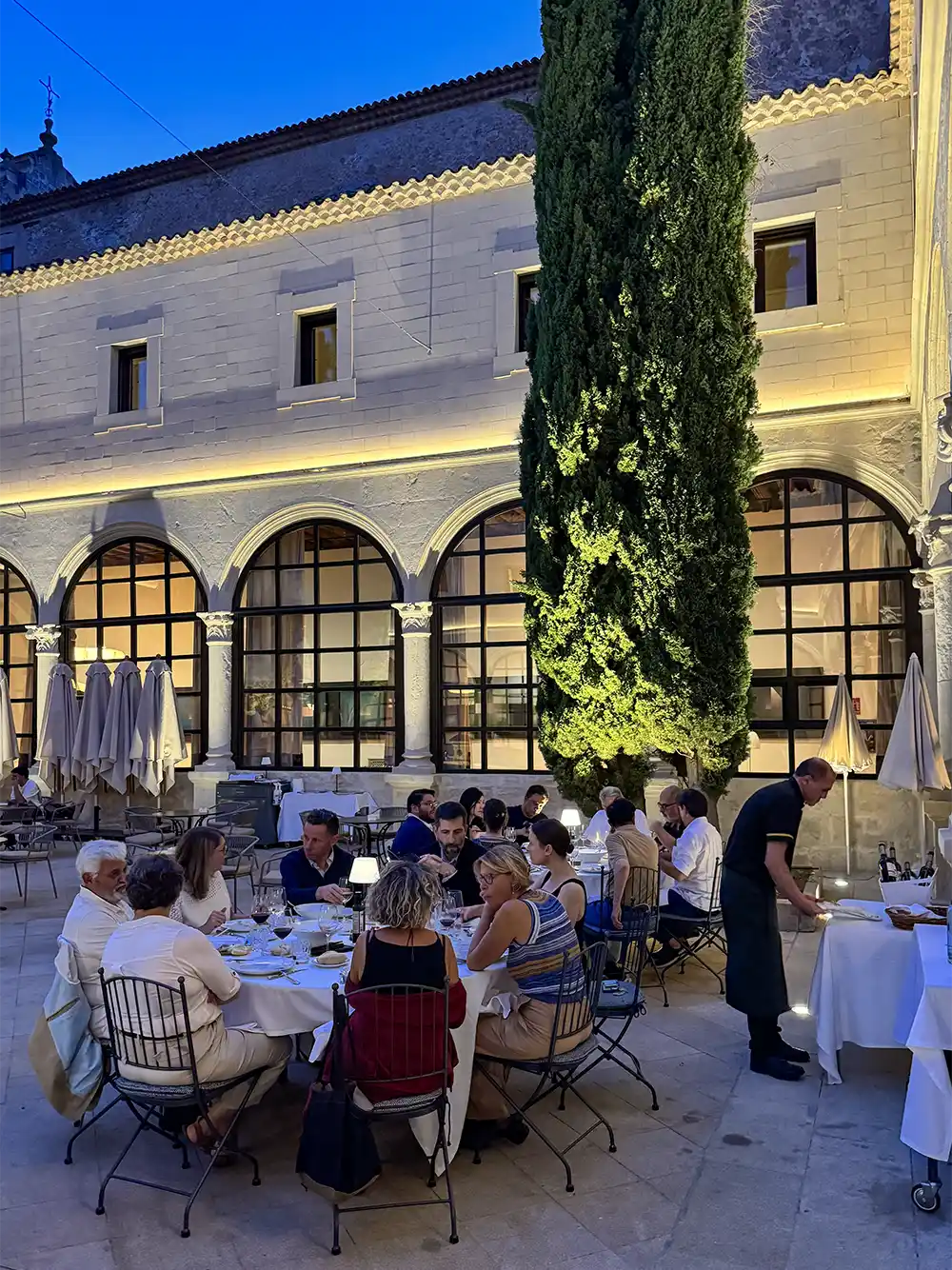 Cuenca-Restaurant-Parador Menschen, die abends im Freien in einem Innenhof mit Bogenfenstern und hohen Bäumen unter warmer Beleuchtung zu Abend essen.