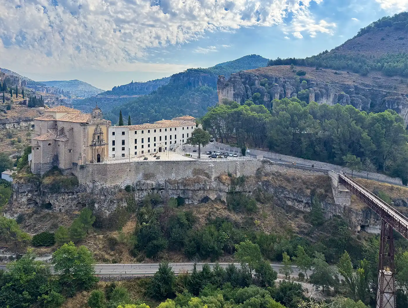 Cuenca-San-Pablo-Parador Ein historisches Kloster aus Stein auf einem Hügel, umgeben von Bäumen, Felsen und einer Brücke an einem teilweise bewölkten Tag.