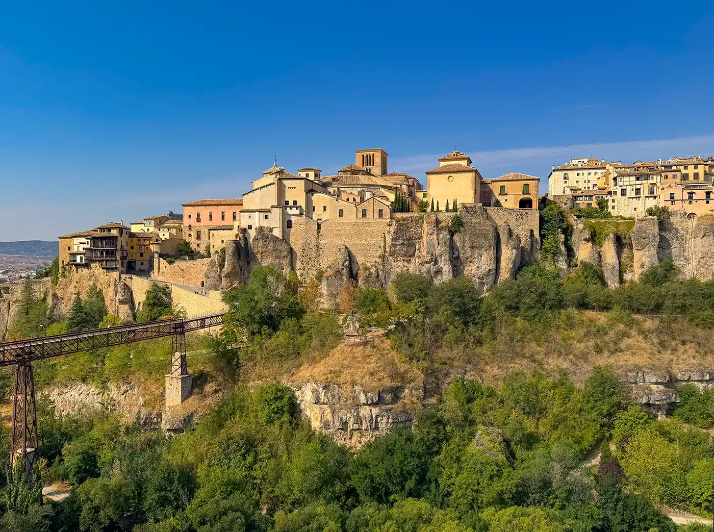 Cuenca-Schlucht-Bruecke Historische Gebäude auf einer felsigen Klippe, umgeben von Grün unter einem strahlend blauen Himmel.