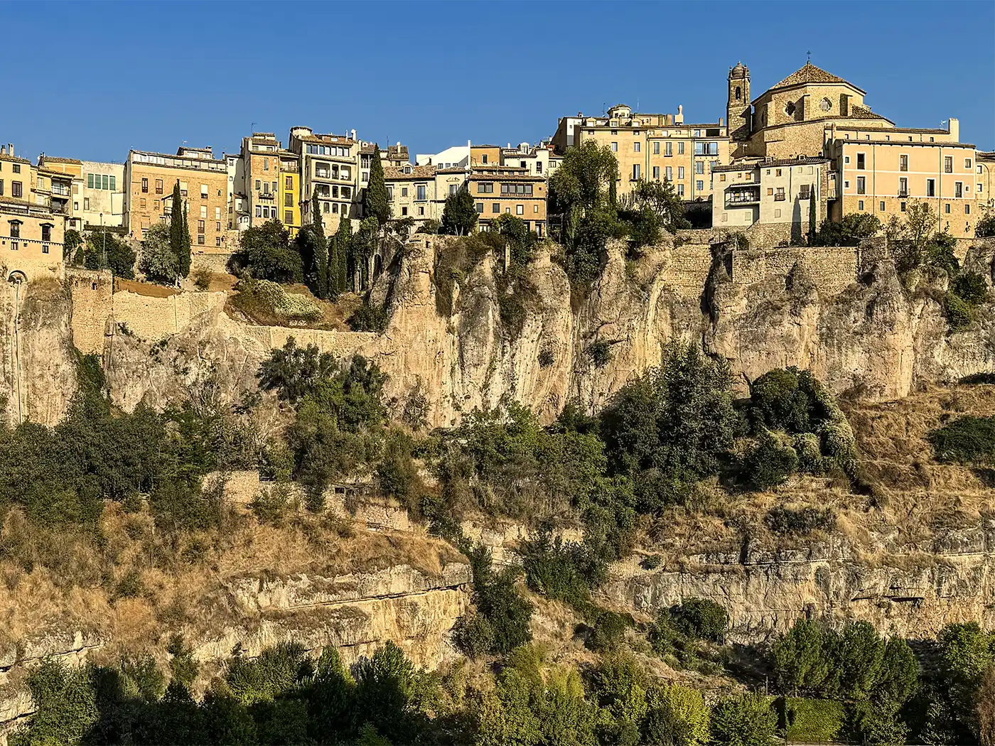 Cuenca-Schlucht-Haeuser Farbenfrohe Gebäude thronen auf felsigen Klippen, umgeben von grüner Vegetation unter einem strahlend blauen Himmel.