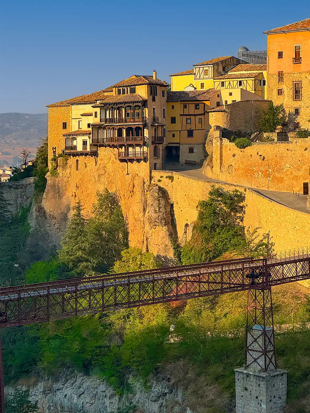 Cuenca-Spanien Farbenfrohe Häuser an der Steilküste mit Holzbalkonen im Sonnenlicht; eine Metallbrücke überspannt die tiefe Schlucht darunter.