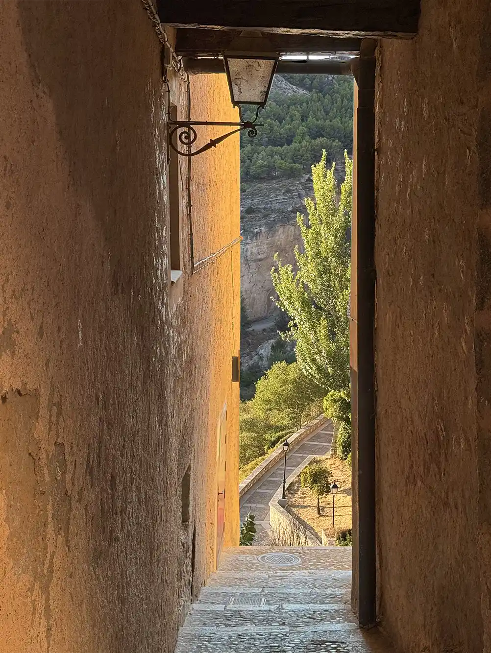 Cuenca-Tordurchgang Eine schmale, von Sonnenlicht durchflutete Steingasse, die den Blick auf Bäume und einen sich darunter schlängelnden Weg freigibt.