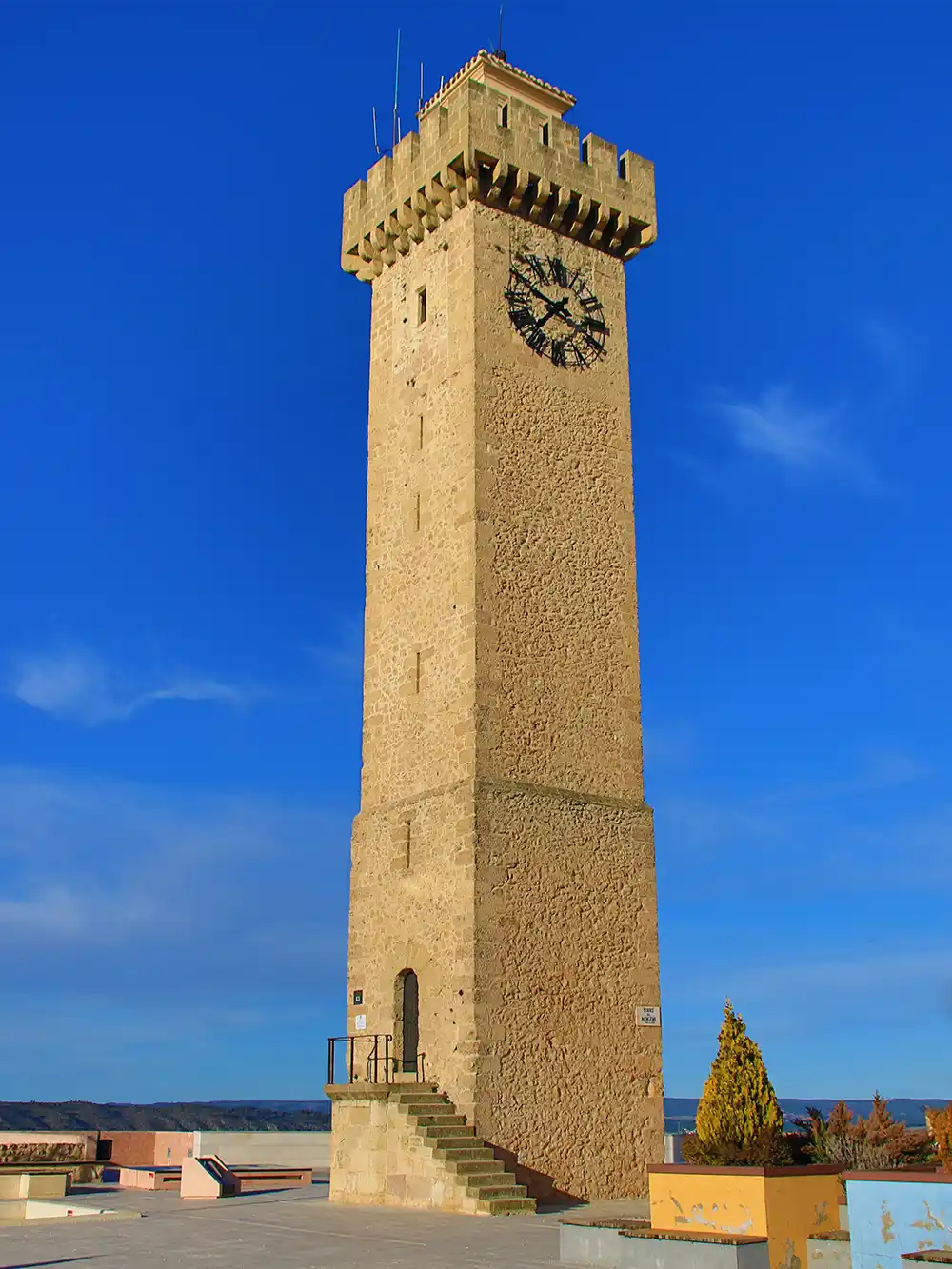 Cuenca-Torre-de-Mangana Ein hoher Uhrturm aus Stein mit Zinnen unter strahlend blauem Himmel, umgeben von einigen niedrigen Gebäuden und Bäumen.