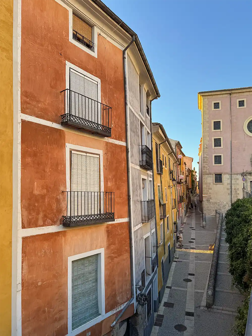 Cuenca-farbige-Haeuser Hohe, farbenfrohe Gebäude säumen eine schmale, sonnige Straße mit einer Treppe und blauem Himmel darüber.