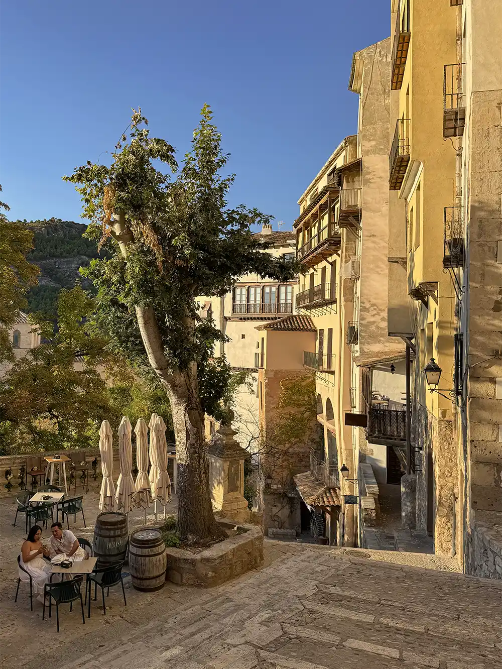 Cuenca-steile-Treppen Ein Paar sitzt an einem Tisch auf einem sonnendurchfluteten Platz neben gelben Gebäuden und Bäumen an einer abfallenden Straße.
