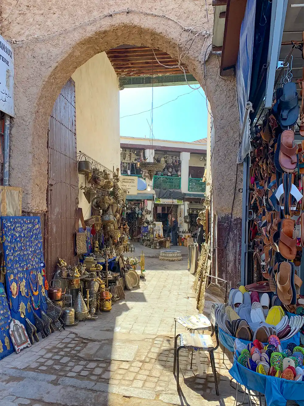 Eingang-Basar Eine schmale Marktstraße, in der unter einem steinernen Torbogen an den Wänden bunte Schuhe, Sandalen und Metallwaren ausgestellt sind.