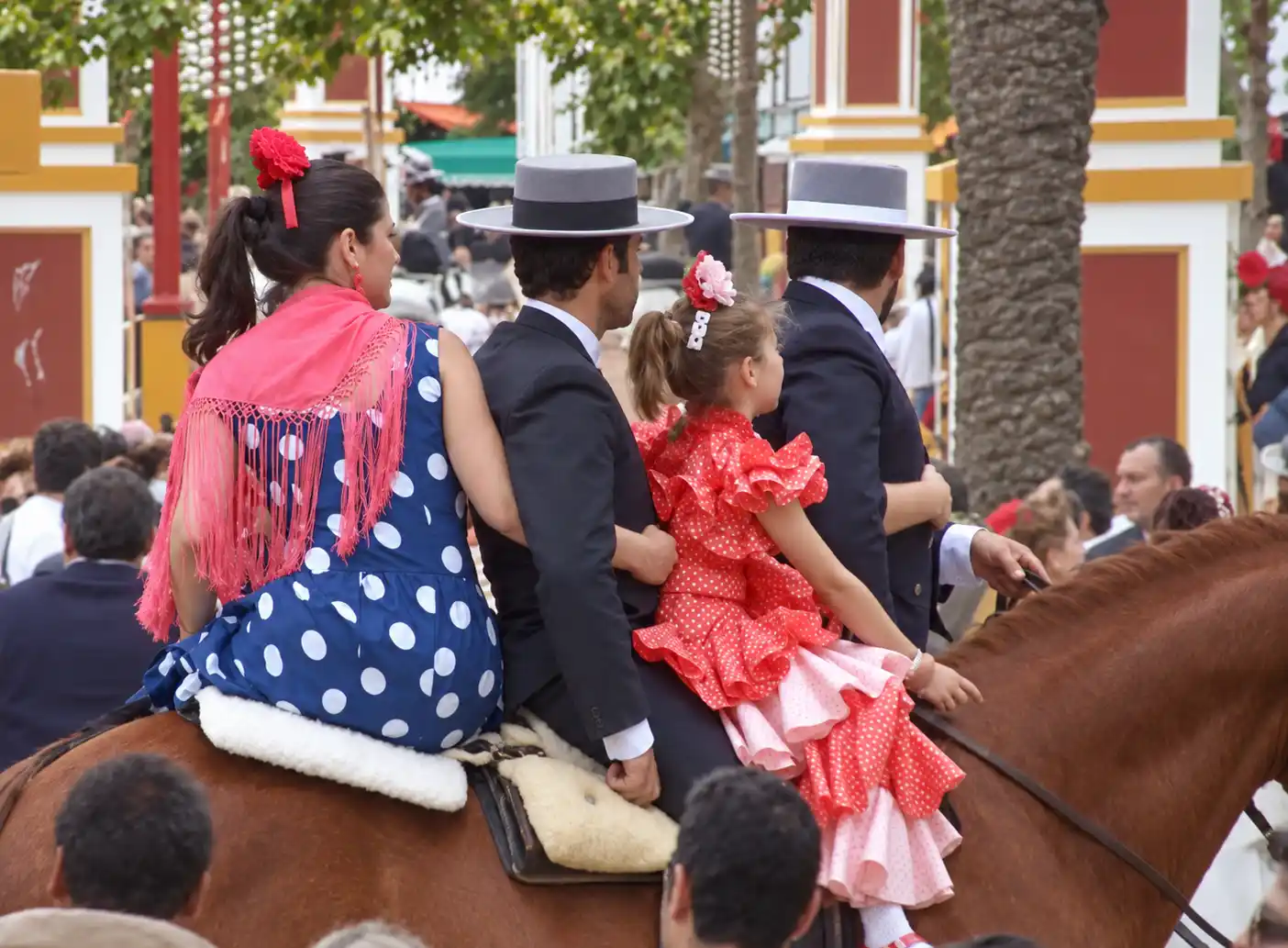 Feria-del-Caballo-Familie Vier Personen in traditioneller spanischer Tracht reiten bei einer festlichen Veranstaltung im Freien auf einem braunen Pferd.