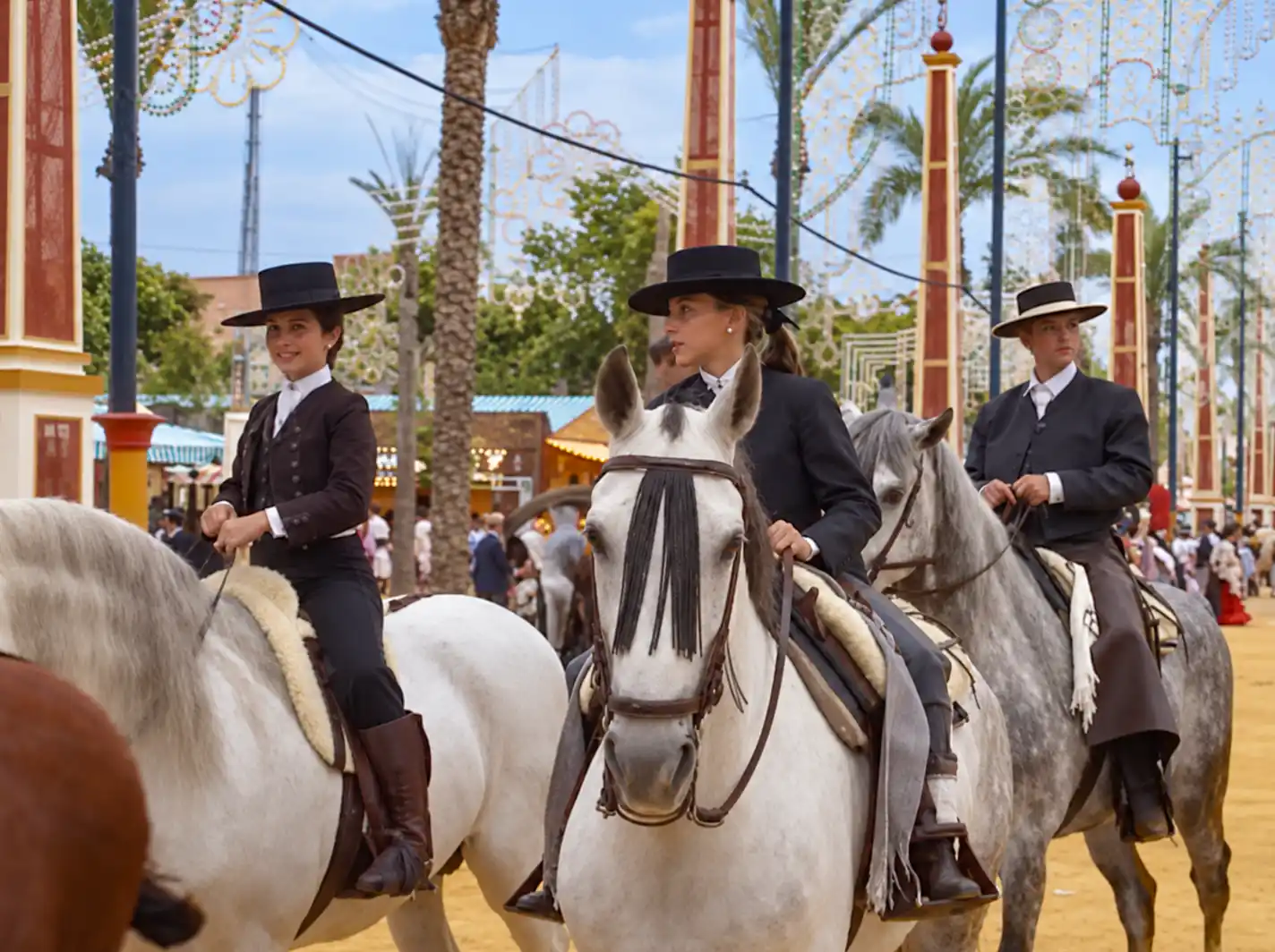 Feria-del-Caballo-Jugendliche Drei Personen in traditioneller Tracht, die auf weißen Pferden reiten, bei einer festlichen Veranstaltung im Freien mit Palmen.