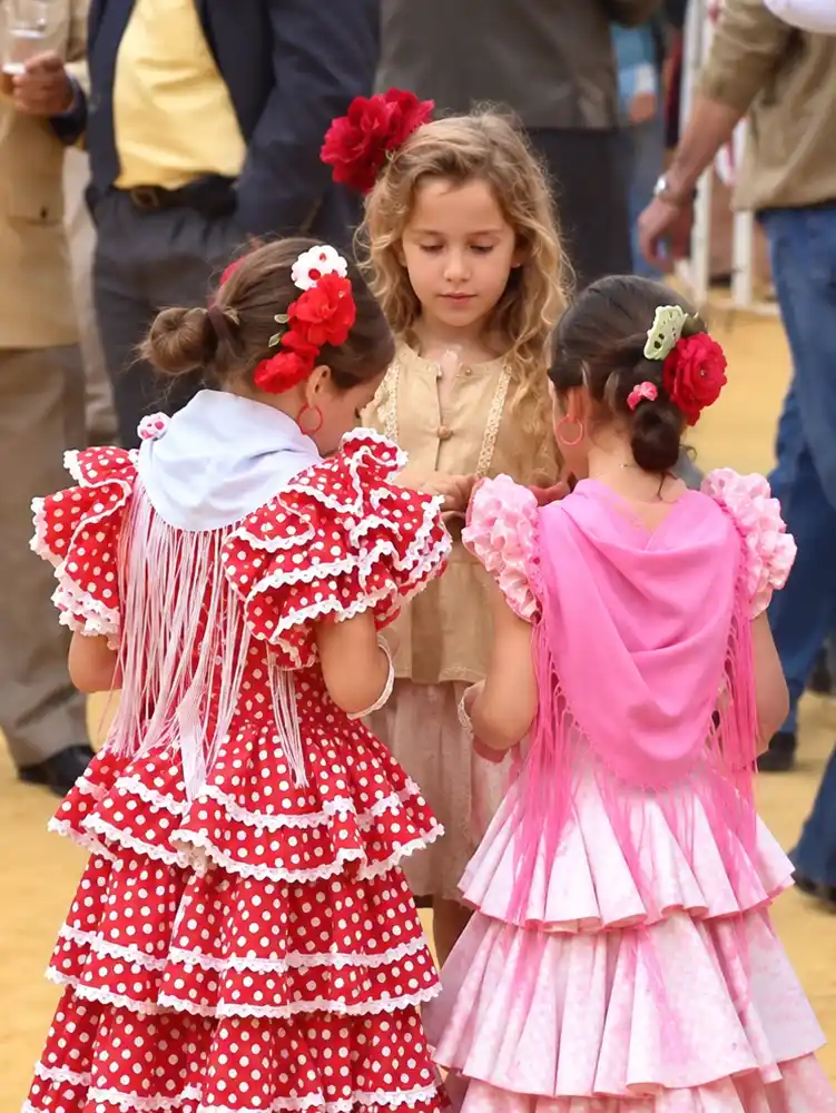 Feria-del-Caballo-Kinder Drei junge Mädchen in traditionellen spanischen Trachten stehen nebeneinander, zwei davon in roten und rosa Rüschen und mit Blumen im Haar.