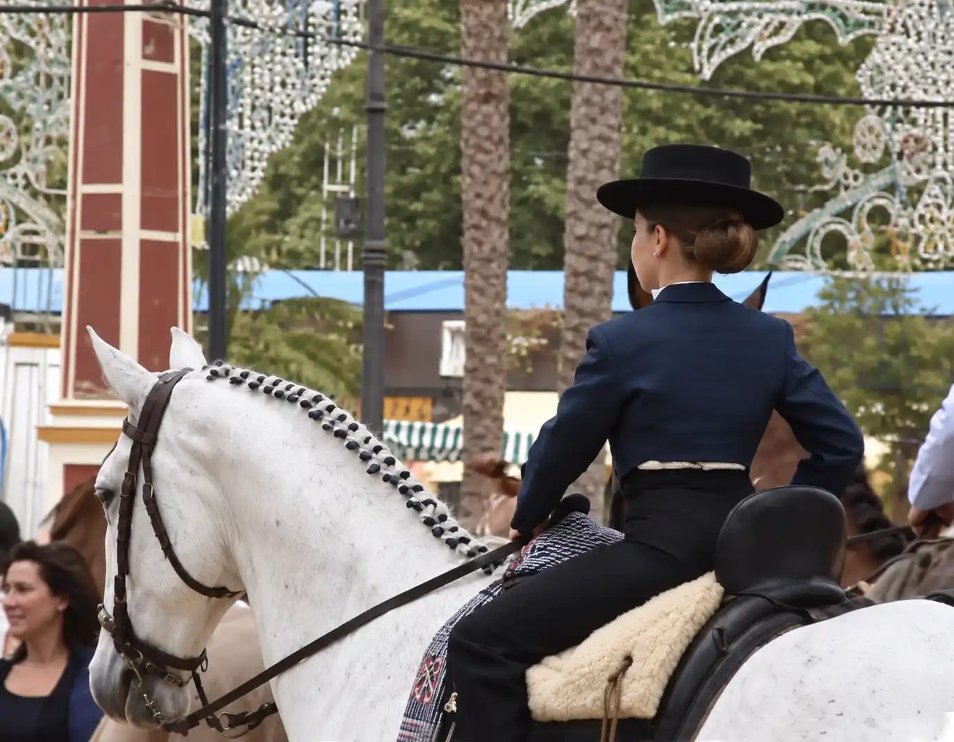 Feria-del-Caballo-Maedchen Eine Frau in traditioneller Tracht reitet auf einem weißen Pferd während einer Veranstaltung im Freien, im Hintergrund sind Bäume und Lichter zu sehen.
