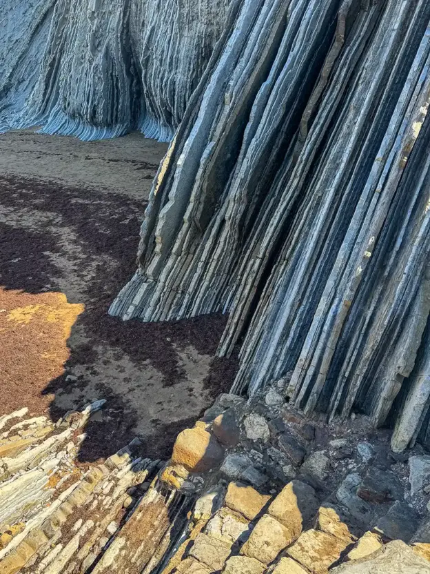 Hohe, geschichtete Felsformationen ragen neben einem Sandstrand empor, auf dem sich hier und da Seetang und Sonnenstrahlen zeigen.