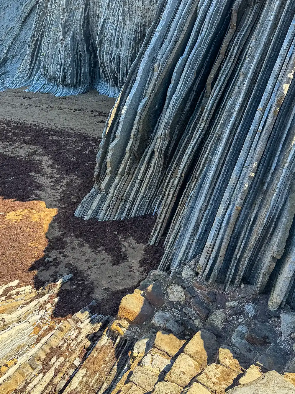 Hohe, geschichtete Felsformationen ragen neben einem Sandstrand empor, auf dem sich hier und da Seetang und Sonnenstrahlen zeigen.