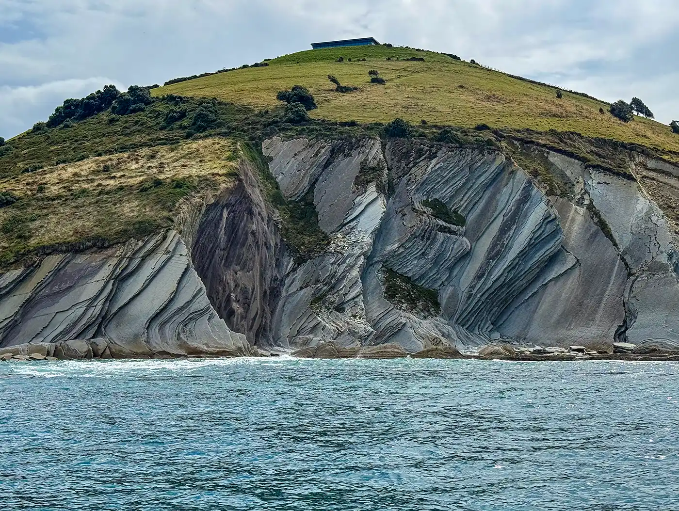 Ein grüner Hügel mit geschichteten Felsklippen, die sich über dem Meer erheben, unter einem bewölkten Himmel.
